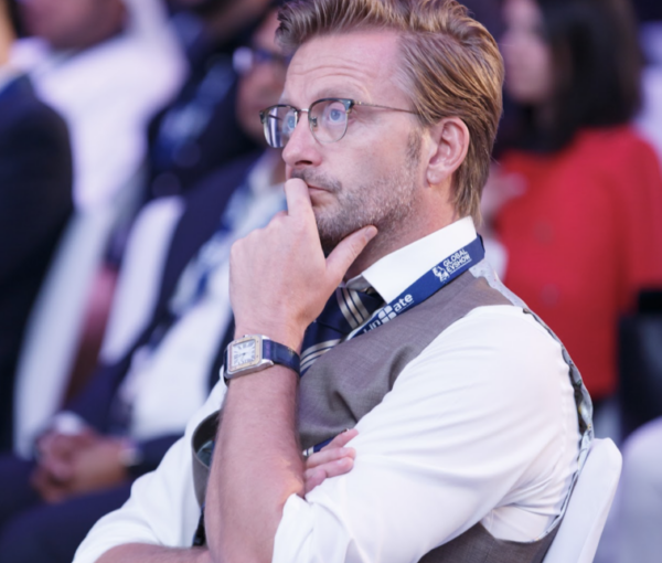 A professionally dressed man with glasses, resting his chin on his hand in a thoughtful pose, amidst a diverse audience at a conference setting.