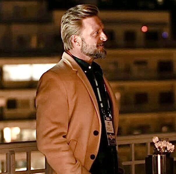 A well-dressed man with a beard stands at a table outdoors, surrounded by blurred city lights in the background.
